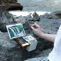 a woman is painting on rocks by the water