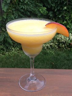 a glass filled with an orange drink on top of a wooden table next to bushes