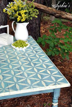 a blue table with flowers and a white pitcher on it in front of a tree