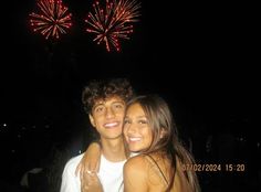 a man and woman standing next to each other with fireworks in the sky behind them