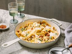 a pasta dish in a white pan on a table next to two glasses and spoons