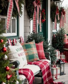 a porch decorated for christmas with red and green plaid pillows, wreaths and trees