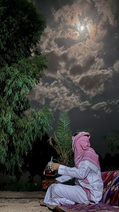 a man sitting on top of a blanket next to a tree under a cloudy sky