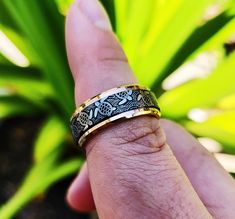 a man's hand with a gold and silver ring on his finger in front of some plants