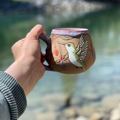 a hand holding a coffee mug with a bird painted on the cup and water in the background