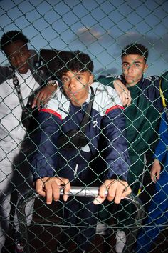 three men standing behind a chain link fence, one holding a cell phone in his hand