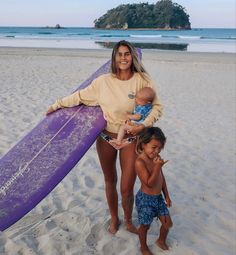 a woman holding a baby and standing next to a purple surfboard on the beach