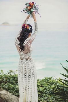 a woman in a long white dress holding flowers up to the sky with her hands