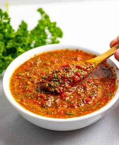 a hand holding a wooden spoon over a bowl of red sauce and garnished with parsley