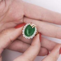 a woman's hand holding a ring with an oval green stone surrounded by diamonds