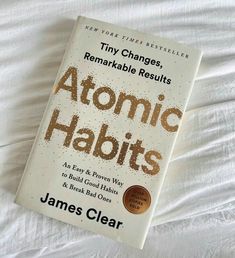 the book atomic habitts by james clear is laying on a white bed sheet and it's title reads tiny changes, remarkable results