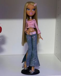 a doll with long blonde hair wearing jeans and a pink top is standing in front of a white wall