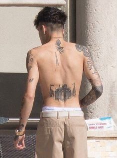 a man with tattoos on his back standing in front of a building and looking down at the ground