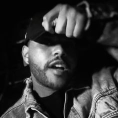 black and white photograph of a man wearing a denim jacket, hat and cap with his hands on his head