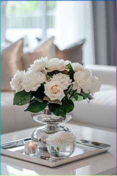 white flowers are in a silver vase on a mirrored tray with candles and pillows behind it