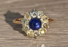 The Tana Antique Mixed Metal Sapphire Cabochon and Old Mine Cut Diamond Halo Ring. Crafted in 14 karat yellow gold and silver, this ring centers on a 1.50 carat royal blue cabochon sapphire. Surrounding the center cabochon sapphire is 1 carat total weight of chunky old mine cut diamonds in a round halo.  The diamonds have a clarity grade of VS2-I1 and a color grade ranging from O-P-Fancy Yellow.  This ring is a ladies finger size 7.5 and is able to be resized to any finger size. Each piece has b Yellow Gold Multi-stone Cabochons For Fine Jewelry, Yellow Gold Multi-stone Cabochons Fine Jewelry, Yellow Gold Diamond Cabochons Fine Jewelry, Vintage Yellow Gold Gemstone Cabochons, Vintage Yellow Gold Cabochons As Gift, Vintage Yellow Gold Cabochons For Gift, Elegant Yellow Gold Cabochons For Wedding, Vintage Gold Sapphire Ring With Cabochon, Vintage Gold Sapphire Cabochon Ring