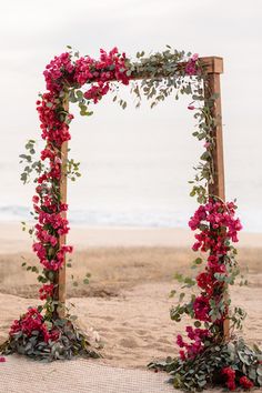 a wooden frame decorated with flowers and greenery on the sand at the beach in front of the ocean