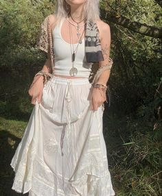 a woman with long white hair wearing a skirt and necklace standing in front of trees