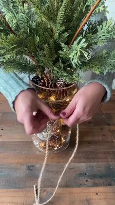 a person holding a glass filled with pine cones and greenery on top of a wooden table