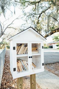 a white book case with books in it sitting on top of a wooden post next to a tree