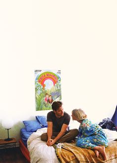 a man and woman sitting on top of a bed in a room with white walls