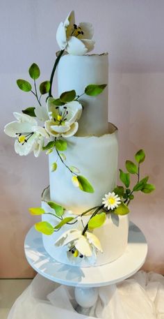 a three tiered cake with white flowers and green leaves on the top, sitting on a plate