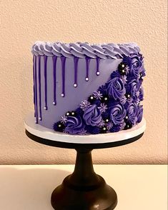 a purple cake with white icing and flowers on a stand in front of a wall