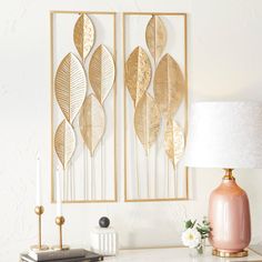 two gold leaf wall hangings on a white wall next to a table with a lamp