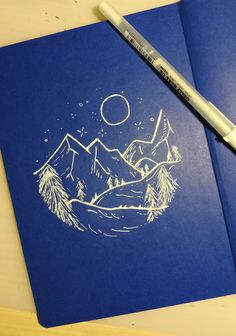 a blue notebook with a drawing of mountains and pine trees on it, next to a marker pen