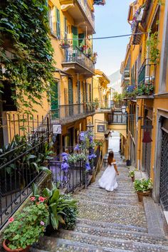 a woman in a white dress is walking down an alley way with flowers on the balconies