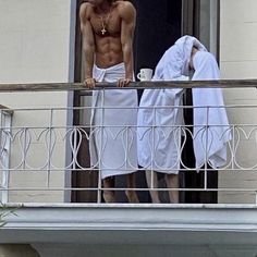 two shirtless men standing on a balcony with towels draped over their heads and looking at each other