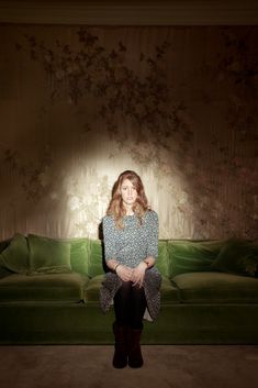 a woman sitting on top of a green couch in front of a wall with flowers