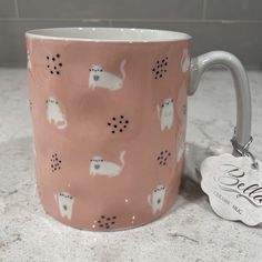 a pink coffee mug with white cats on it and a name tag hanging from the handle