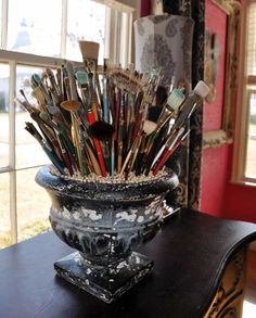 a vase filled with lots of paint brushes on top of a wooden table next to a window