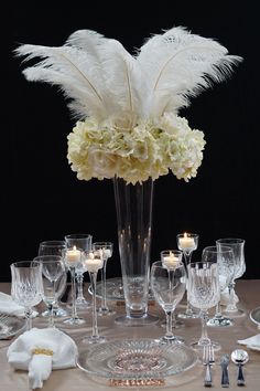 there is a vase with white flowers and feathers on it, surrounded by wine glasses