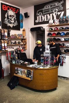 two men standing at a counter in a store with hats on it and skateboards behind them