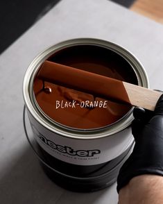 a person holding a paint brush over a can of black - orange colored paint with the word written on it