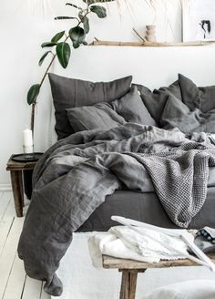 an unmade bed with grey linens in a white room next to a potted plant