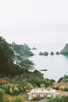 an outdoor table set up on the side of a cliff overlooking water and land with rocks in the background