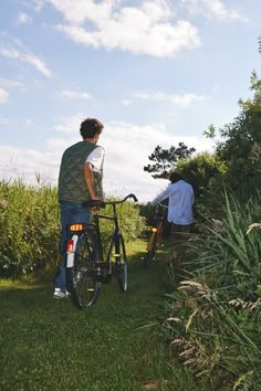 two men standing next to each other with their bikes on the grass and bushes behind them