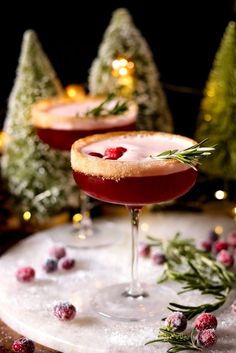 two glasses filled with red liquid and garnished with greenery, surrounded by small christmas trees