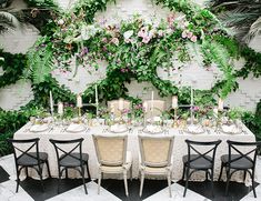 a table set up with white linens and greenery on the wall behind it