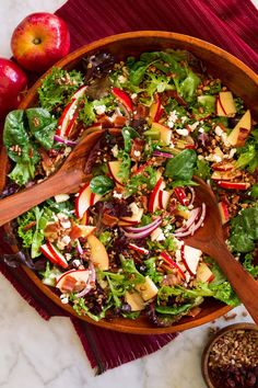 a salad in a bowl with wooden spoons and apples on the table next to it