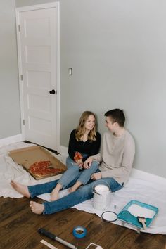 a man and woman sitting on the floor with pizza in front of them, looking at each other