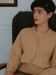 a woman sitting in a chair wearing a brown sweater and black hat with pearls on her ear