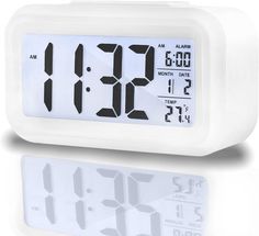 the alarm clock is white and has black numbers on its face, while it sits in front of a reflective surface