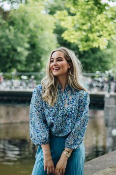 a woman standing in front of a river wearing a blue floral shirt and pleated skirt