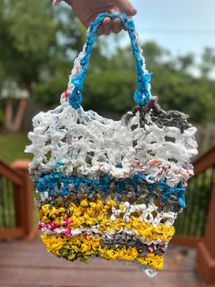 a hand holding a multicolored bag made from plastic bottles and string, in front of a wooden deck