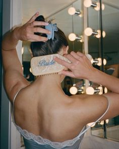 a woman brushes her hair in front of a mirror with lights on the wall behind her