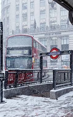 a red double decker bus driving down a snow covered street next to a sidewalk sign